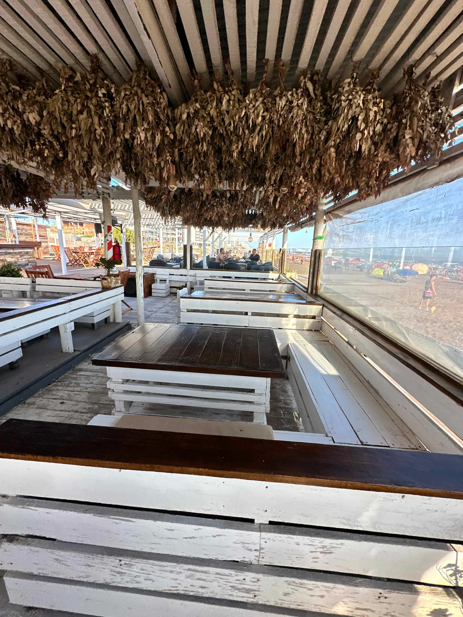 Mesa de madera y bancos blancos bajo la pérgola de SHANTI, con la playa a un costado, en un día de verano.